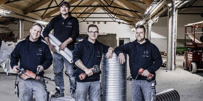 Die Gruppe von vier Männern in Arbeitskleidung posiert mit verschiedenen Werkzeugen, während sie in einem Baustellenumfeld stehen.