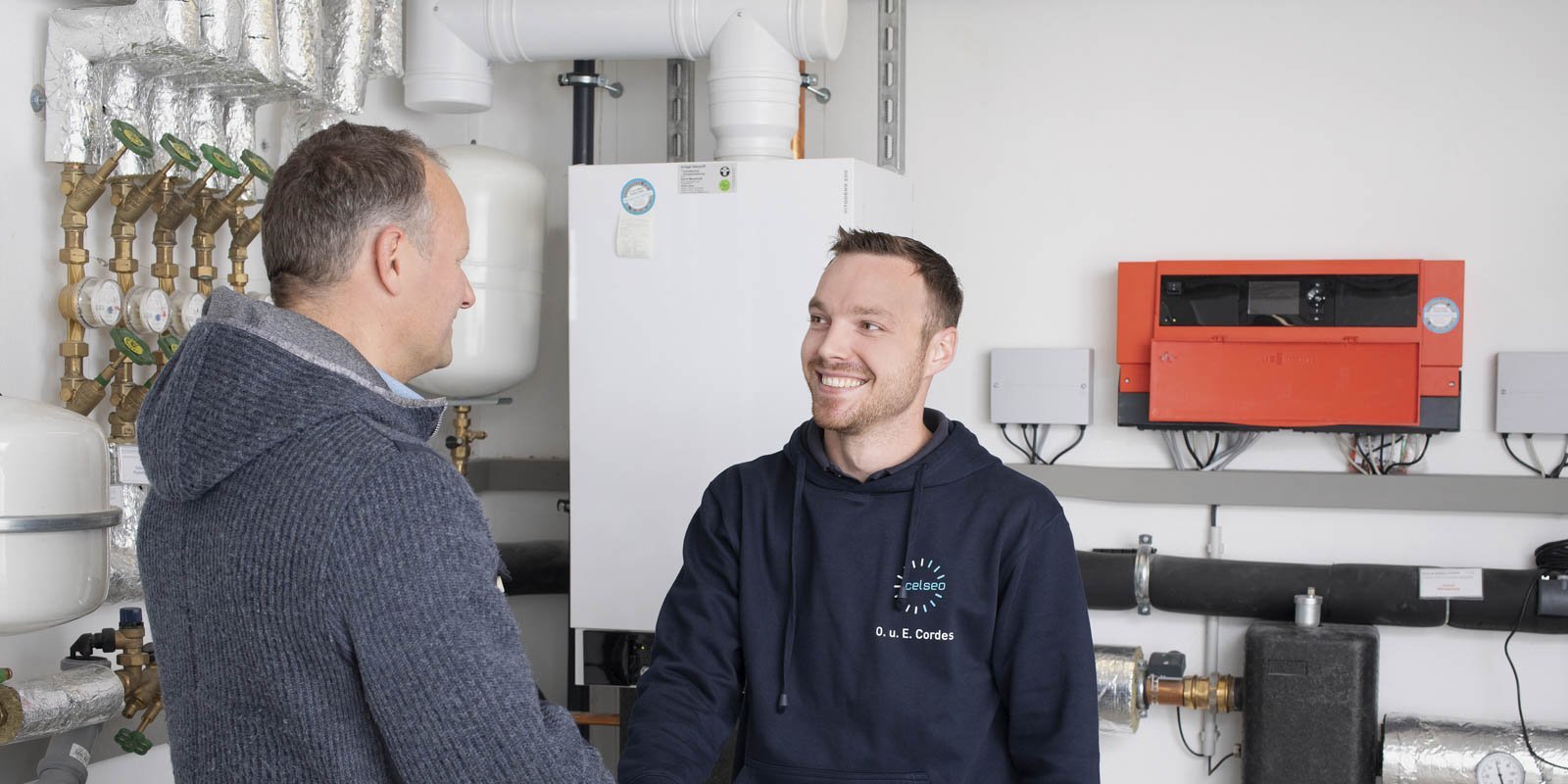 Zwei Männer schütteln sich die Hände in einem modernen Technikraum mit Heizungsanlagen und Rohren im Hintergrund.