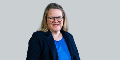 Eine lächelnde Frau mit blonden Haaren trägt eine dunkle Jacke und eine blaue Bluse, vor einem grauen Hintergrund.