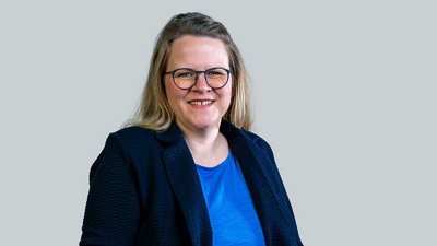 Eine lächelnde Frau mit blonden Haaren trägt eine dunkle Jacke und eine blaue Bluse, vor einem grauen Hintergrund.