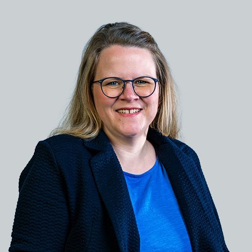 Eine lächelnde Frau mit langen, blonden Haaren trägt eine Brille und einen schwarzen Blazer über einem blauen Oberteil.